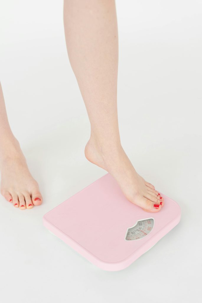 pexels-photo-6975473-6975473 A woman's feet with red nails stepping onto a pink weight scale, symbolizing health and wellness.